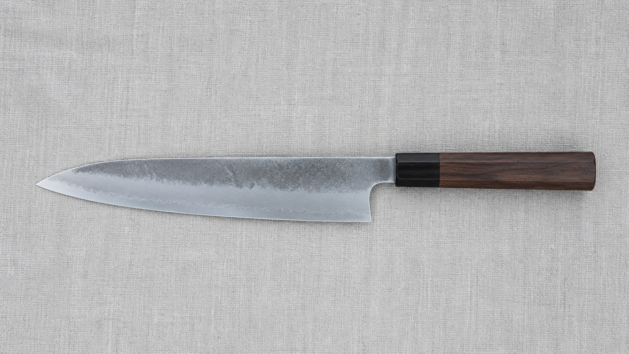 A symmetrical double bevel Japanese gyuto knife laid flat on a light grey linen cloth, photographed from directly above with even diffused lighting showing both sides of the blade edge. No text, no labels, no logos.