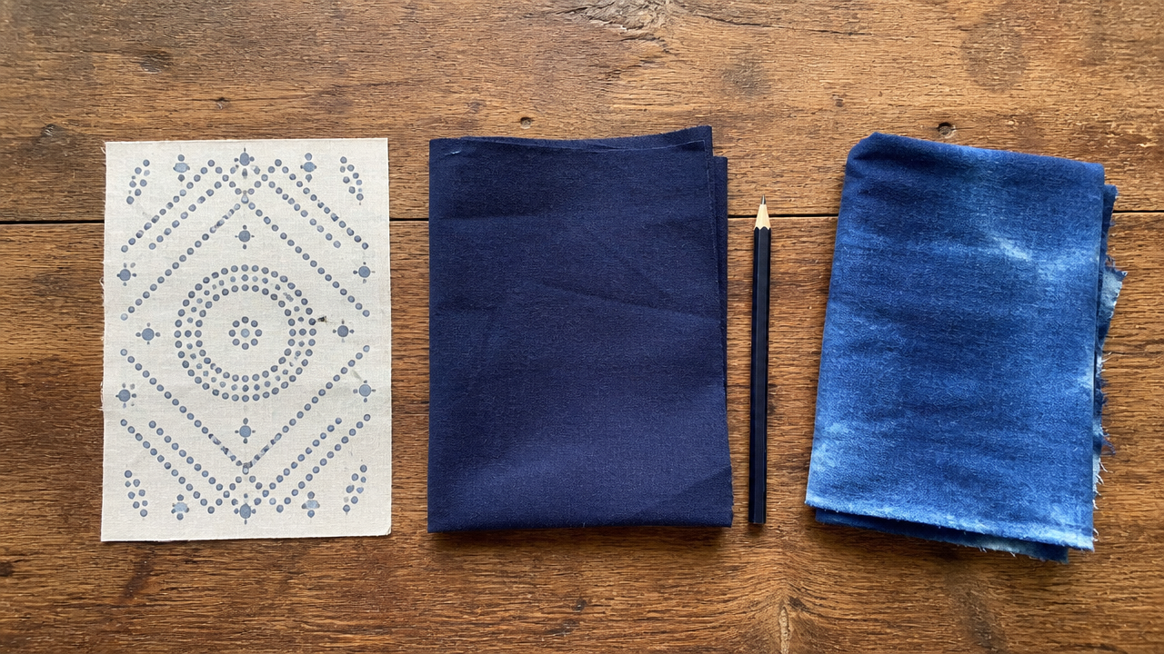 A flat lay showing three distinct craft project stages on a wooden table: a small pre-printed fabric panel with geometric dot patterns, a piece of navy cotton fabric next to a thin pencil, and a folded piece of slightly uneven indigo-dyed cloth. Natural daylight. No text, no brand logos, no stitching shown yet.