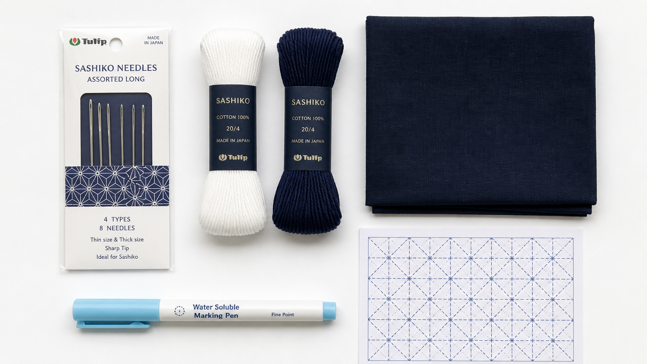 A clean flat-lay on a white background showing essential sashiko beginner supplies: a packet of Tulip sashiko needles, two spools of sashiko thread (one white, one navy), a folded piece of dark indigo cotton fabric, a water-soluble marking pen, and a small printed geometric pattern grid. Soft overhead lighting, minimal shadows, product photography style.