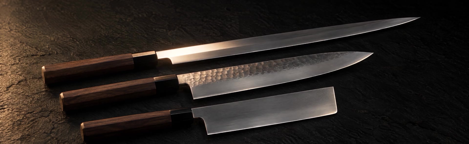 Three Japanese kitchen knives — yanagiba, hammered gyuto and nakiri — on dark slate