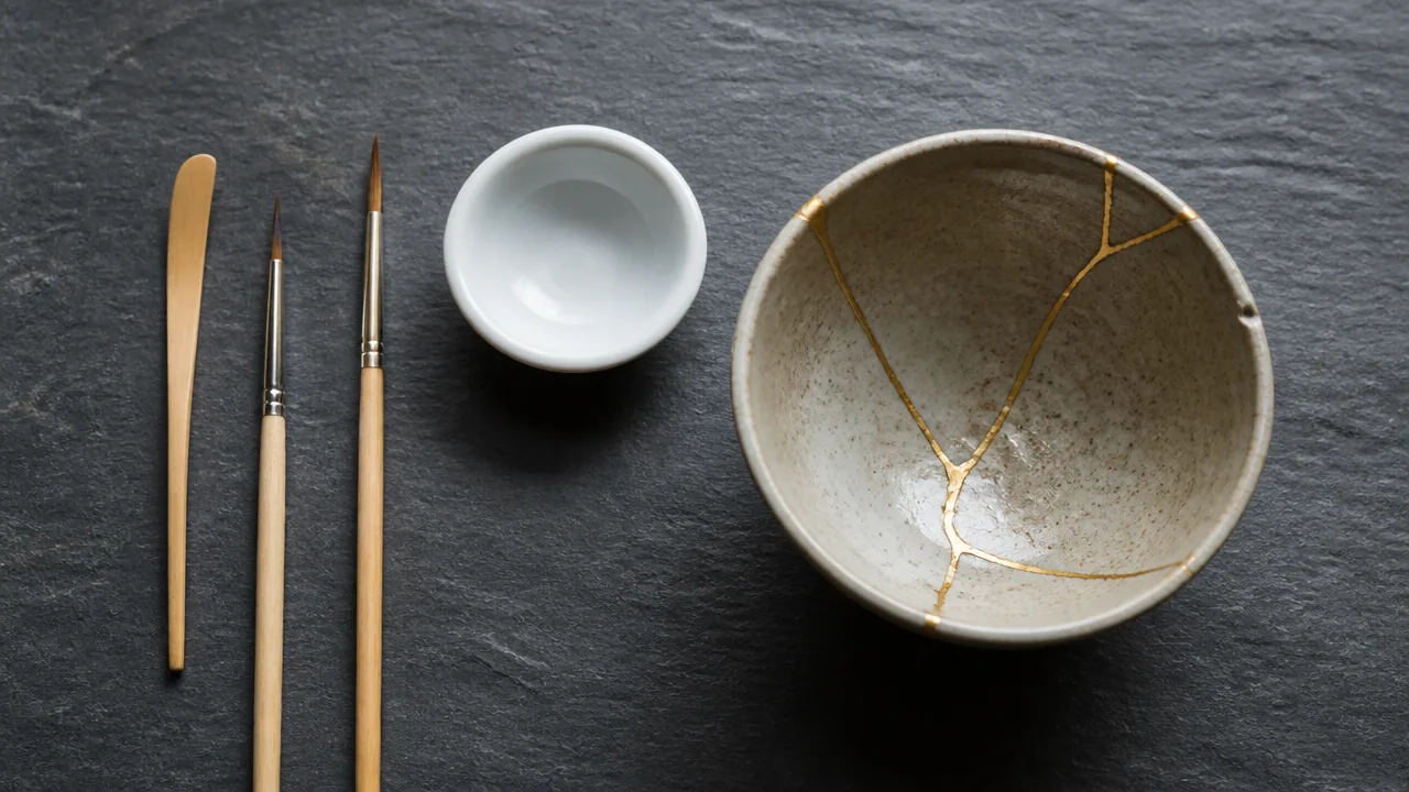 A flat lay photograph on a dark slate surface showing a set of Japanese kintsugi repair tools arranged neatly: a thin bamboo spatula, two small fine-bristled brushes of slightly different sizes, and a small round porcelain mixing dish, alongside a repaired ceramic bowl with visible gold seams. Soft natural window light, no hands, no text, no lettering, no logos, no price tags, photorealistic style.