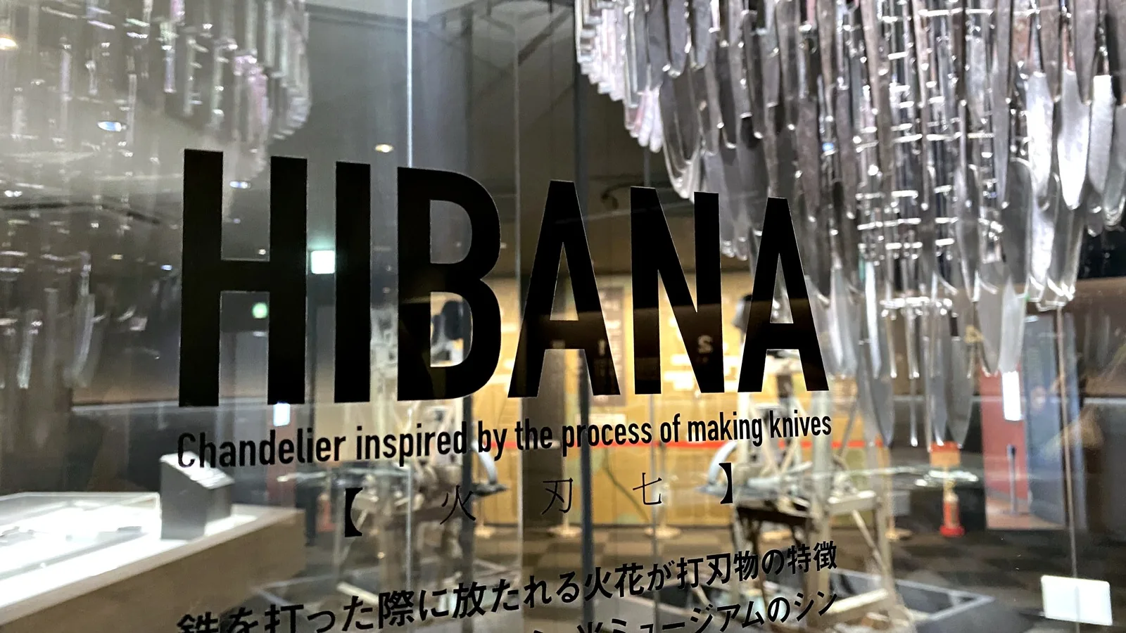 HIBANA sign on the glass case reading Chandelier inspired by the process of making knives, with the knife chandelier behind it