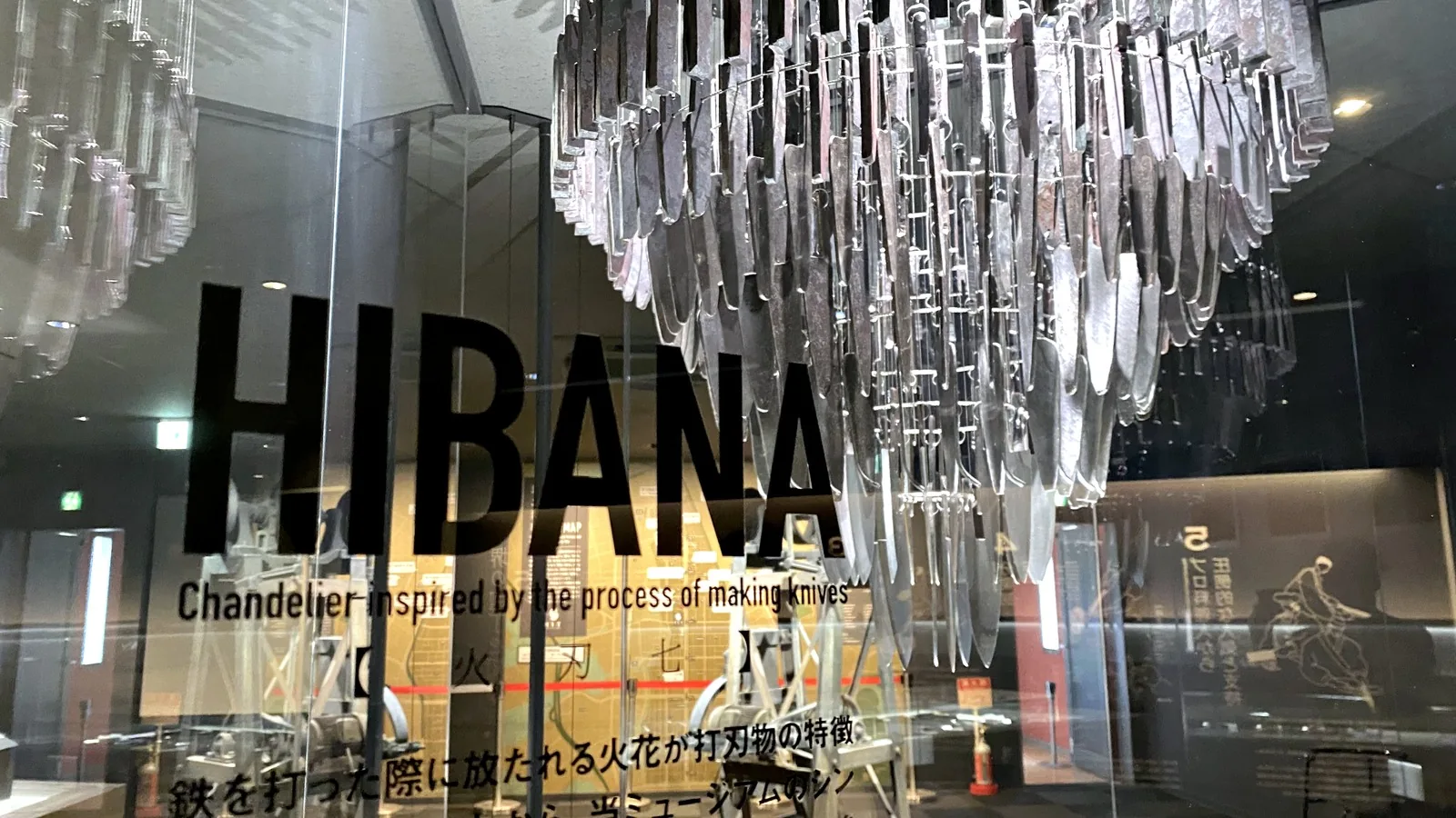 HIBANA sign on the glass enclosure at Sakai Knife Museum CUT, with the subtitle Chandelier inspired by the process of making knives, the characters 火刃七 and a Japanese line about sparks, with the knife chandelier behind it
