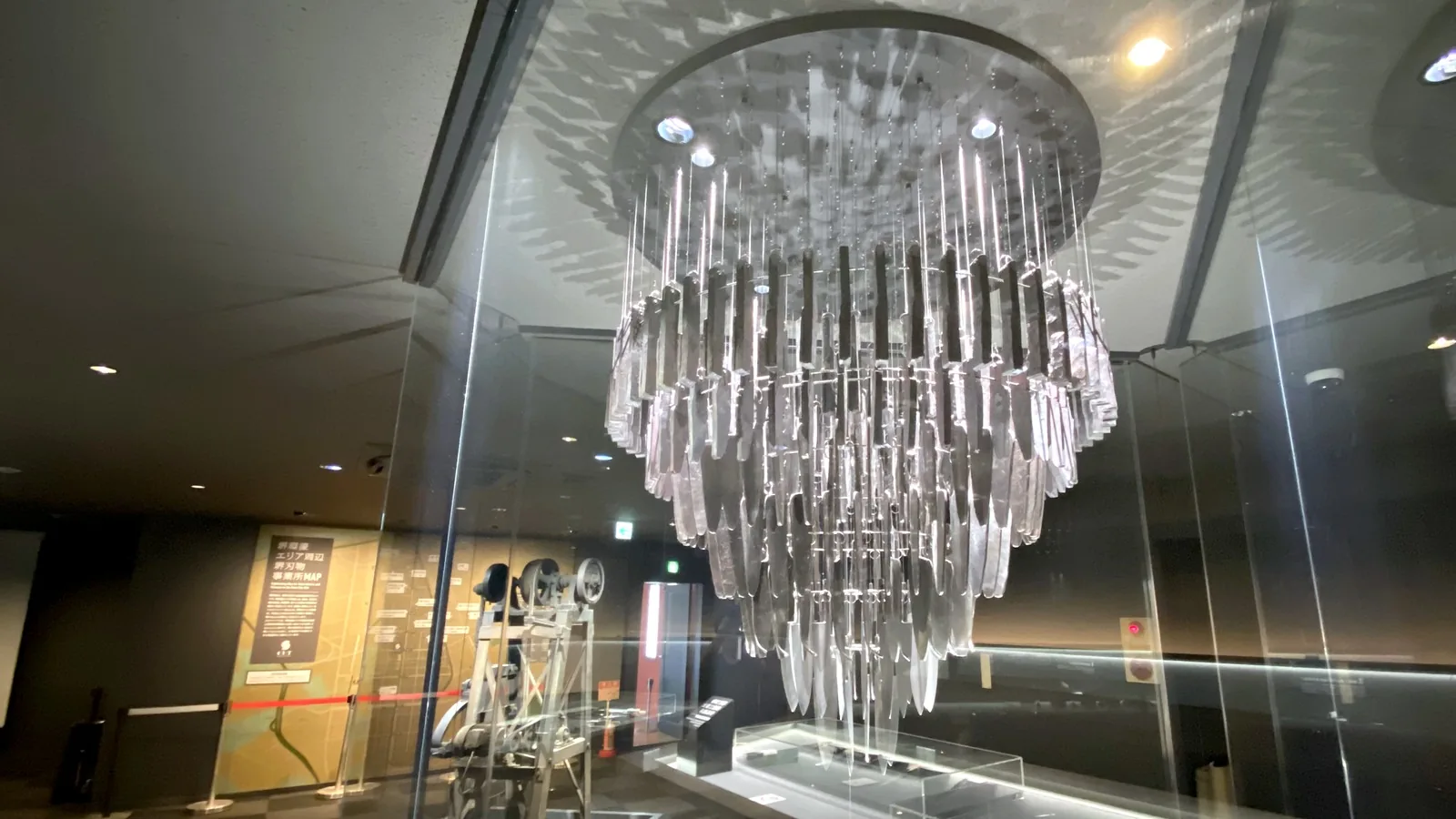 The HIBANA chandelier inside its glass enclosure, with the museum's belt hammer and a wall map of local knife businesses behind it