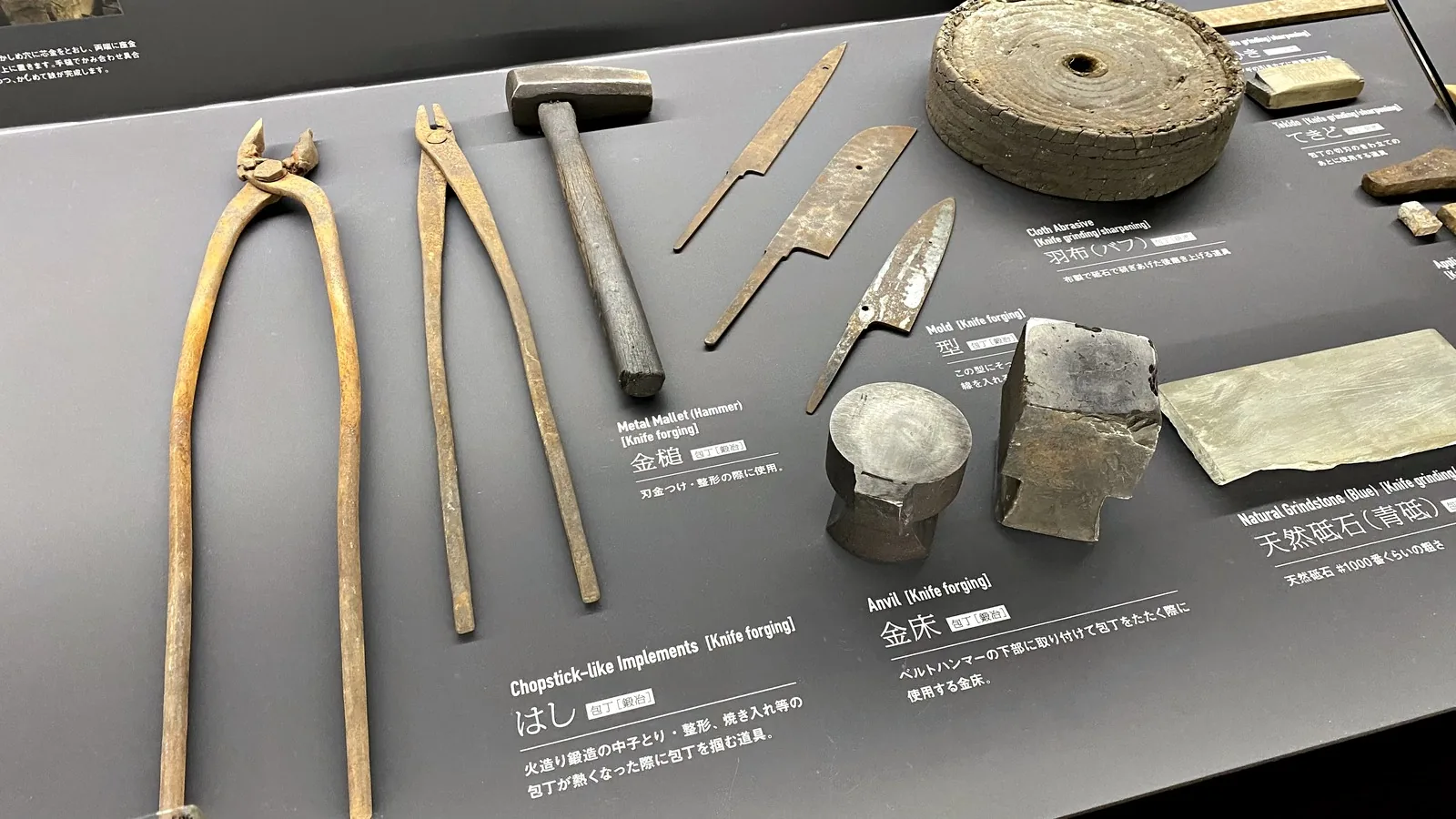 Traditional knife-making tools on display: long tongs, a hammer, an anvil, a natural whetstone and a cloth buffing wheel