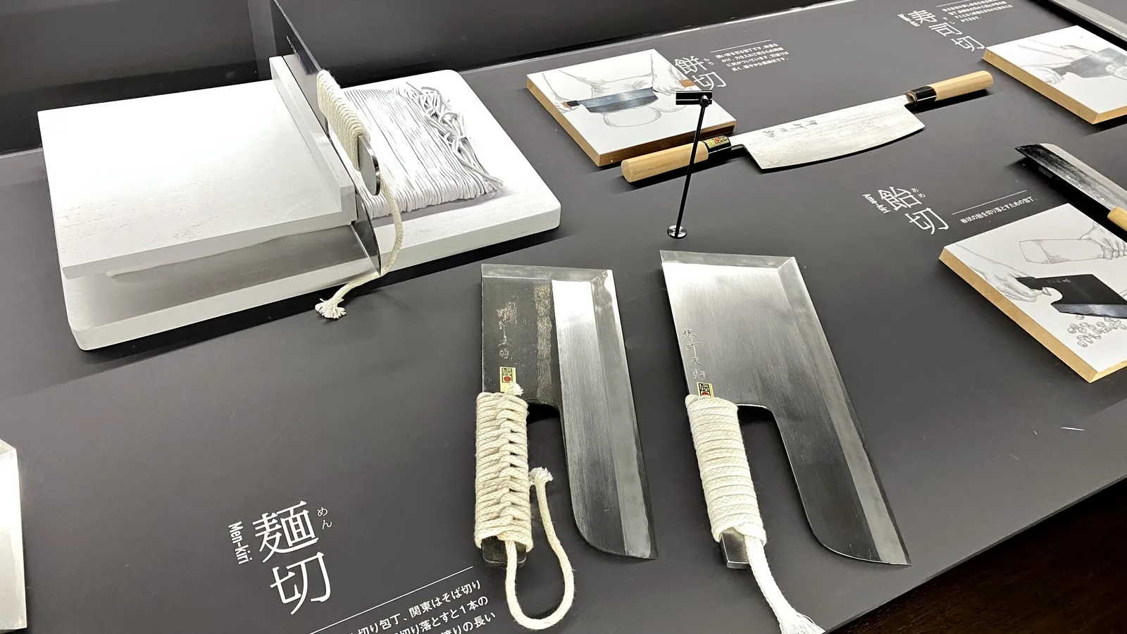 Two menkiri noodle knives with cord-wrapped handles displayed next to a noodle cutting board and guide