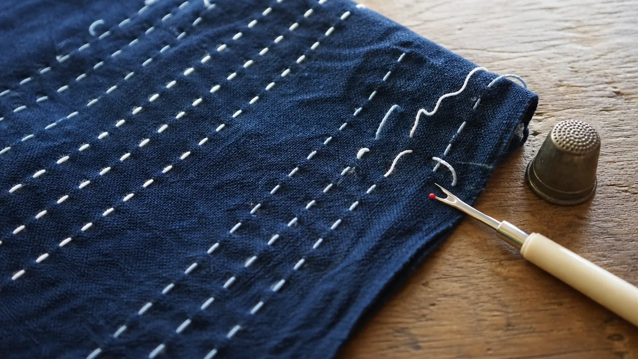 A close-up photograph of a piece of indigo cotton fabric with several neat rows of white running stitches, a few of the stitches at the end of one row partly unpicked, a small seam ripper and a thimble lying beside it on a wooden table. Soft natural window light. Photorealistic photograph, soft natural window light, realistic fabric texture with visible weave and hand stitches, not an illustration or painted rendering. No hands, no fingers, no people, no text, no lettering, no numbers, no logos, no brand names, no price tags.