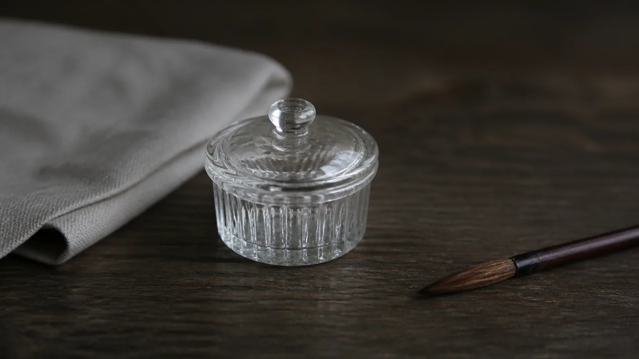 A quiet still life photograph on a dark wooden surface of a single small lidded glass jar and one fine brush lying flat beside a folded linen cloth, generous negative space, low natural light, calm mood. Photorealistic photograph, macro lens, soft natural light, realistic materials with natural texture and subtle reflections, not an illustration or painted rendering. No hands, no people, no text, no lettering, no numbers, no logos, no brand names, no price tags.