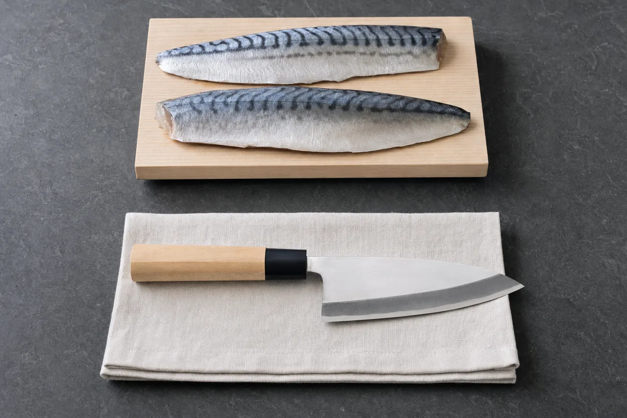 A deba knife on a folded linen cloth below two mackerel fillets on a small wooden board