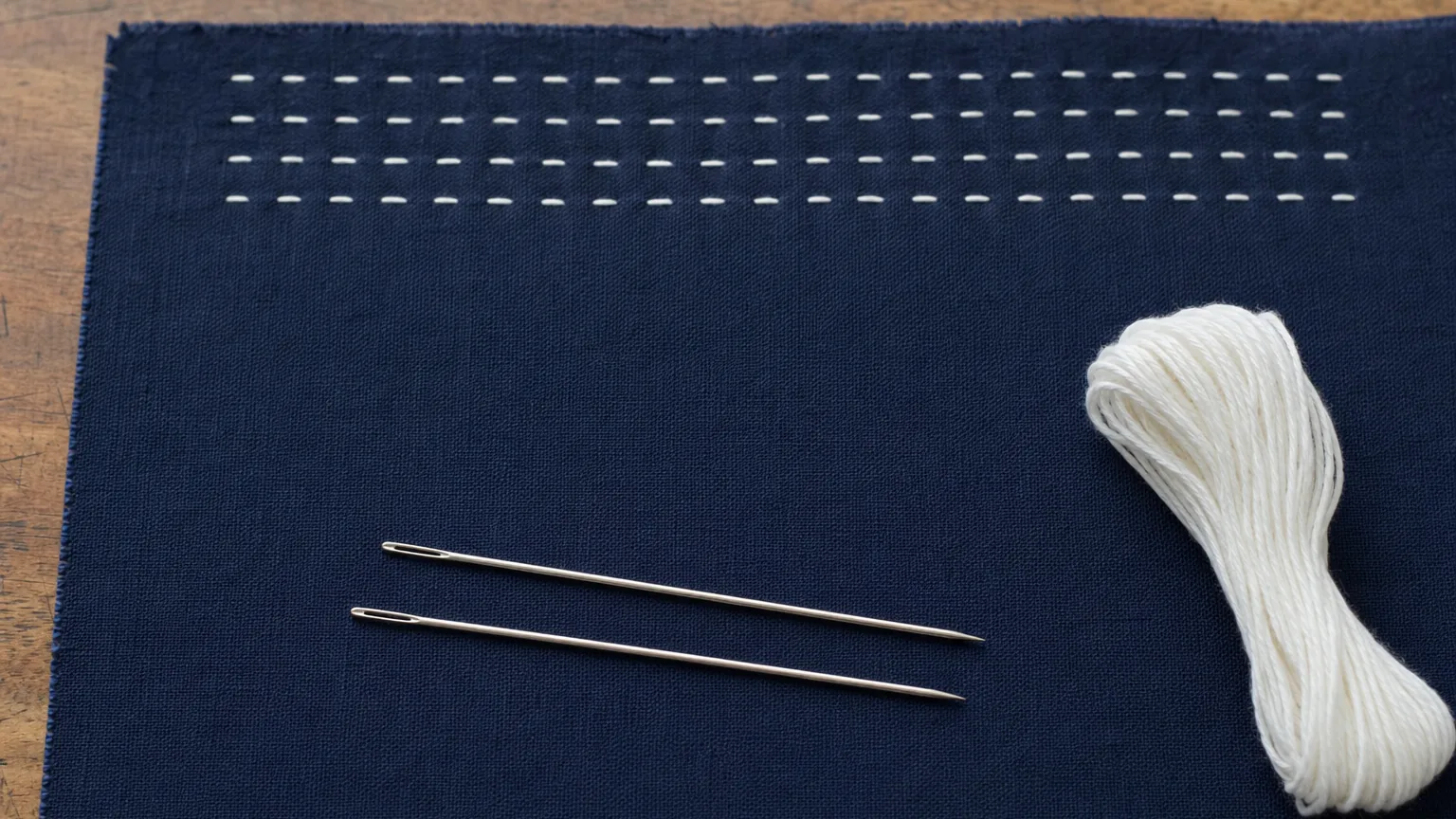Two sashiko needles and a skein of white sashiko thread on indigo cotton cloth with rows of white running stitches