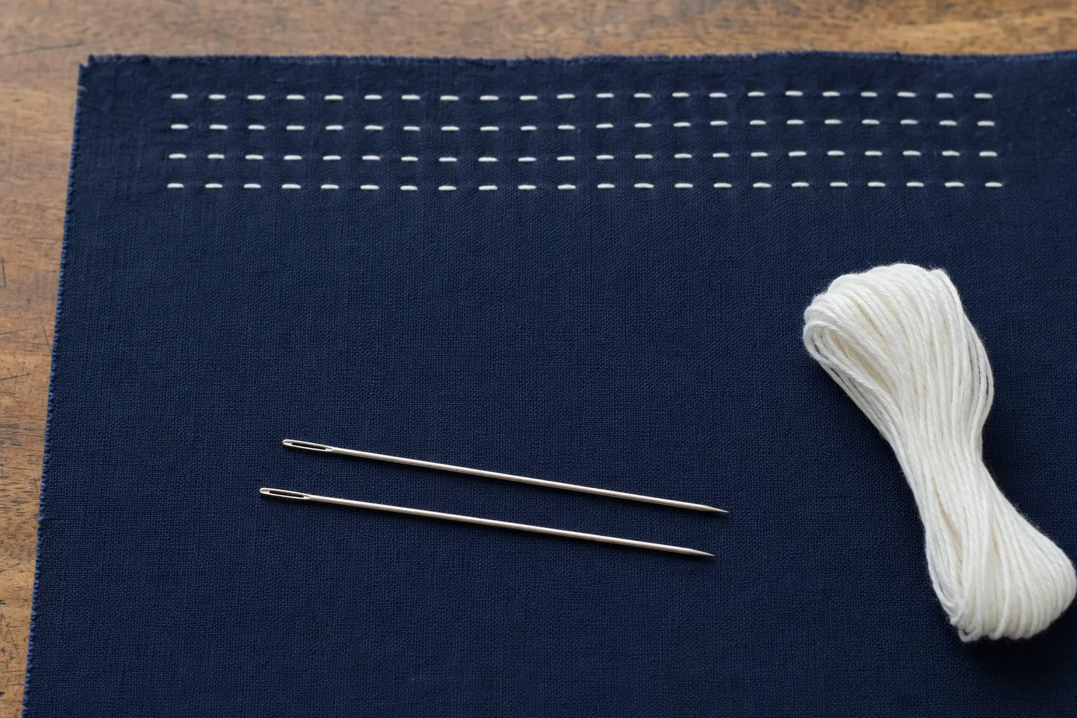 Two unthreaded sashiko needles and a skein of white sashiko thread lying separately on indigo cotton cloth with rows of white running stitches