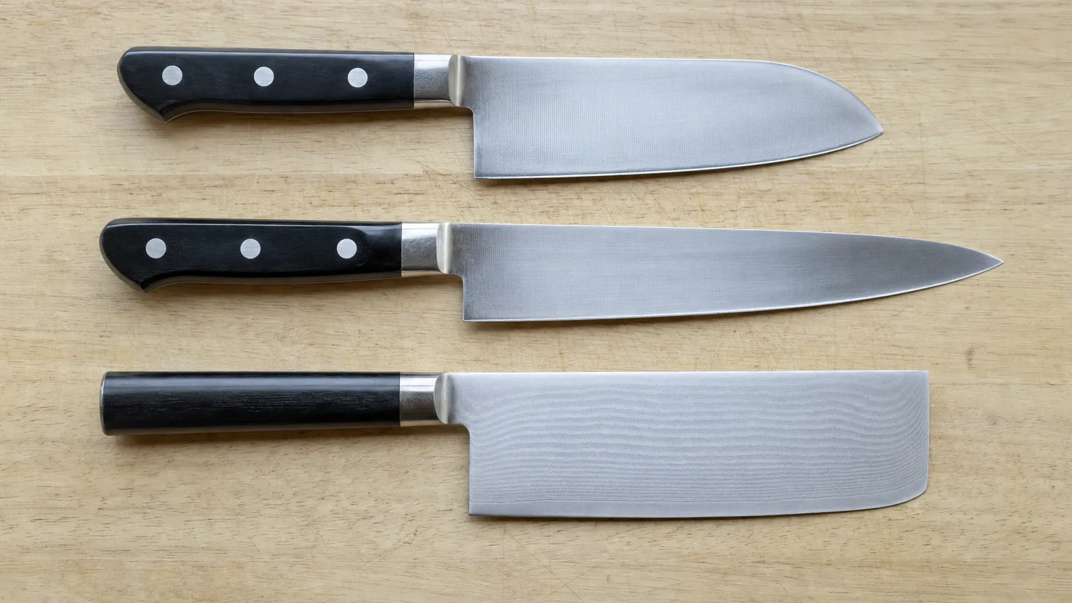Santoku, gyuto and nakiri knives lying in three rows on a wooden cutting board, handles on the left